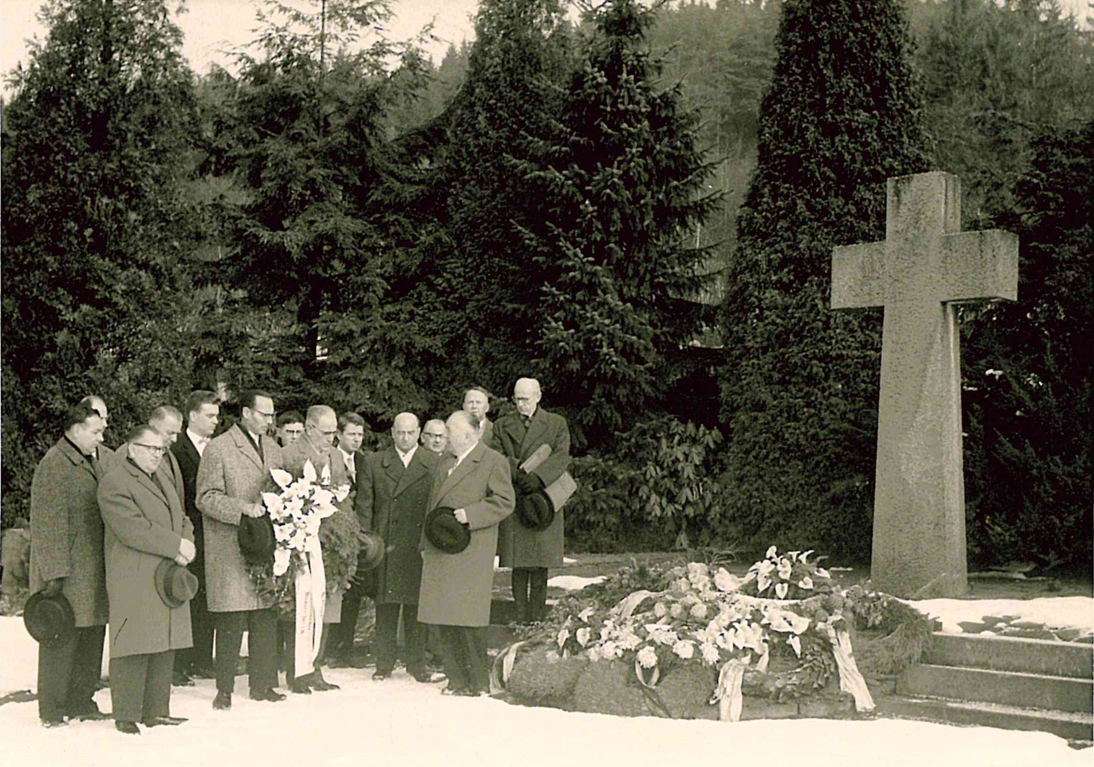 Das Bild zeigt 11 Männer, die vor einem Kreuz, einem Ehrenmal stehen und Kränze niederlegen. 